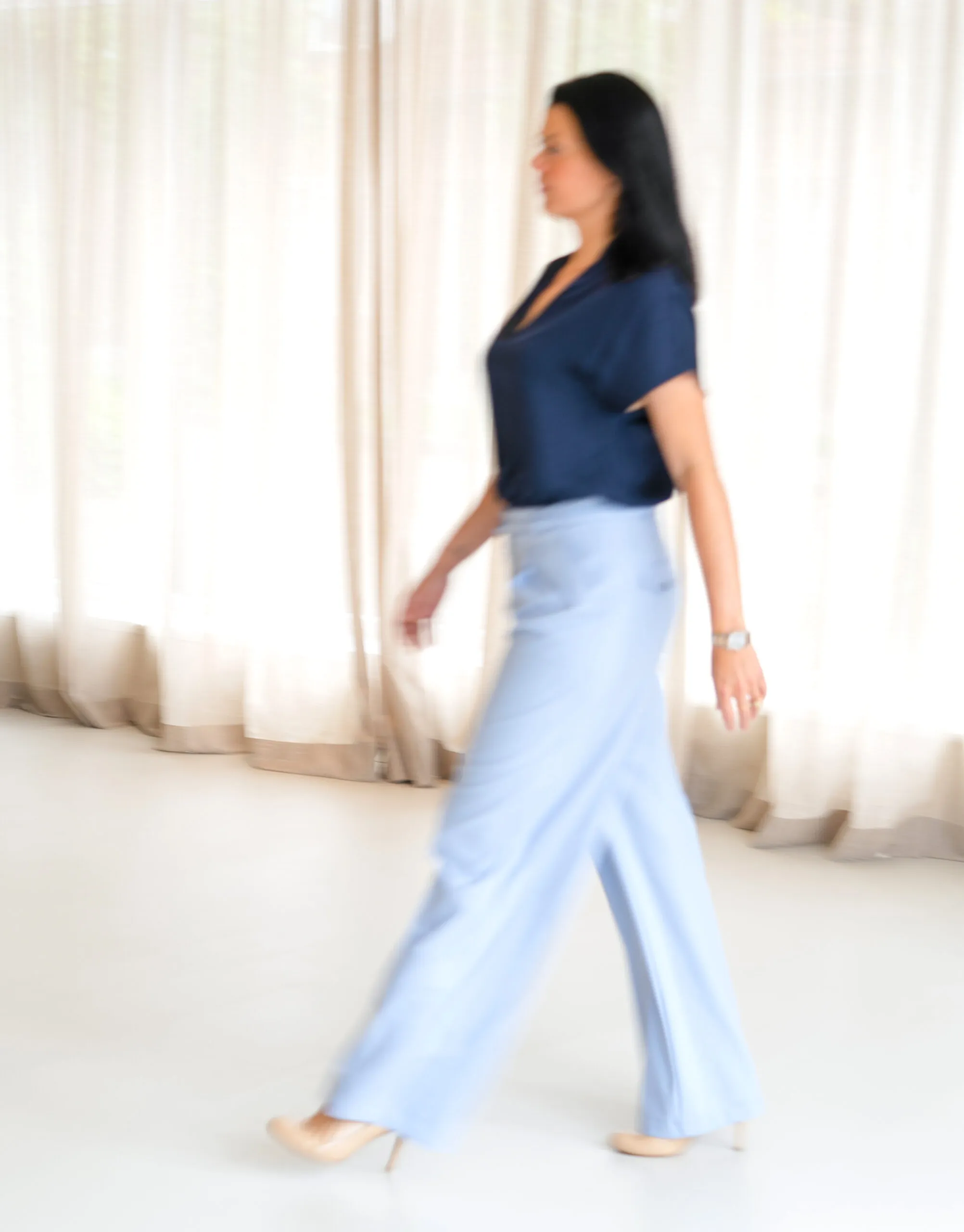 Dineke geht in einer hellblauen Hose und einer dunkelblauen Bluse durch einen hellen Raum mit Vorhängen, aufgenommen mit Bewegungsunschärfe.