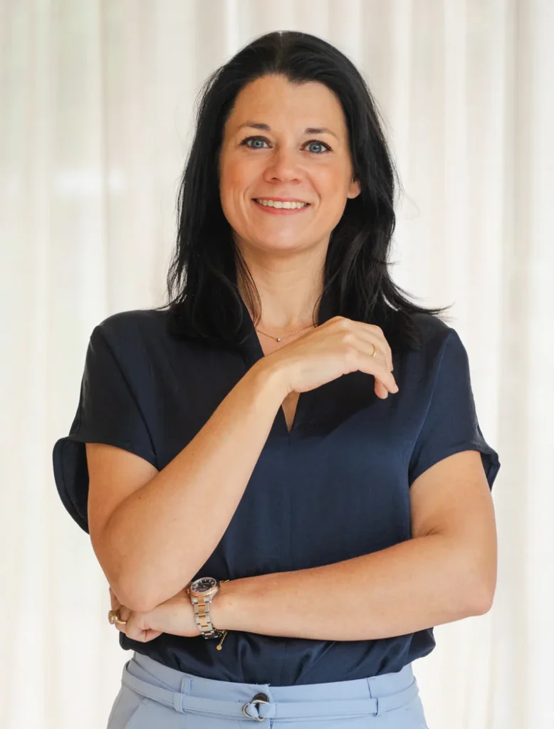 Dineke staat glimlachend met gekruiste armen in een donkerblauwe blouse voor lichte gordijnen.