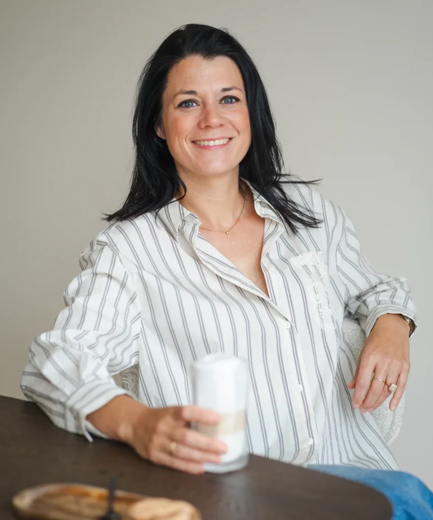 Vrouw in gestreepte blouse glimlacht terwijl ze aan een tafel zit met een glas in haar hand.