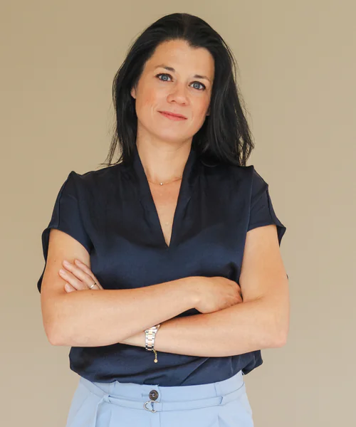 Vrouw met donker haar staat met gekruiste armen en een zelfverzekerde uitstraling voor een neutrale achtergrond.