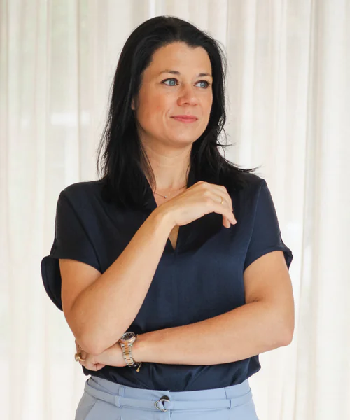 Dineke kijkt opzij met haar hand op haar borst, in een donkerblauwe blouse voor lichte gordijnen.