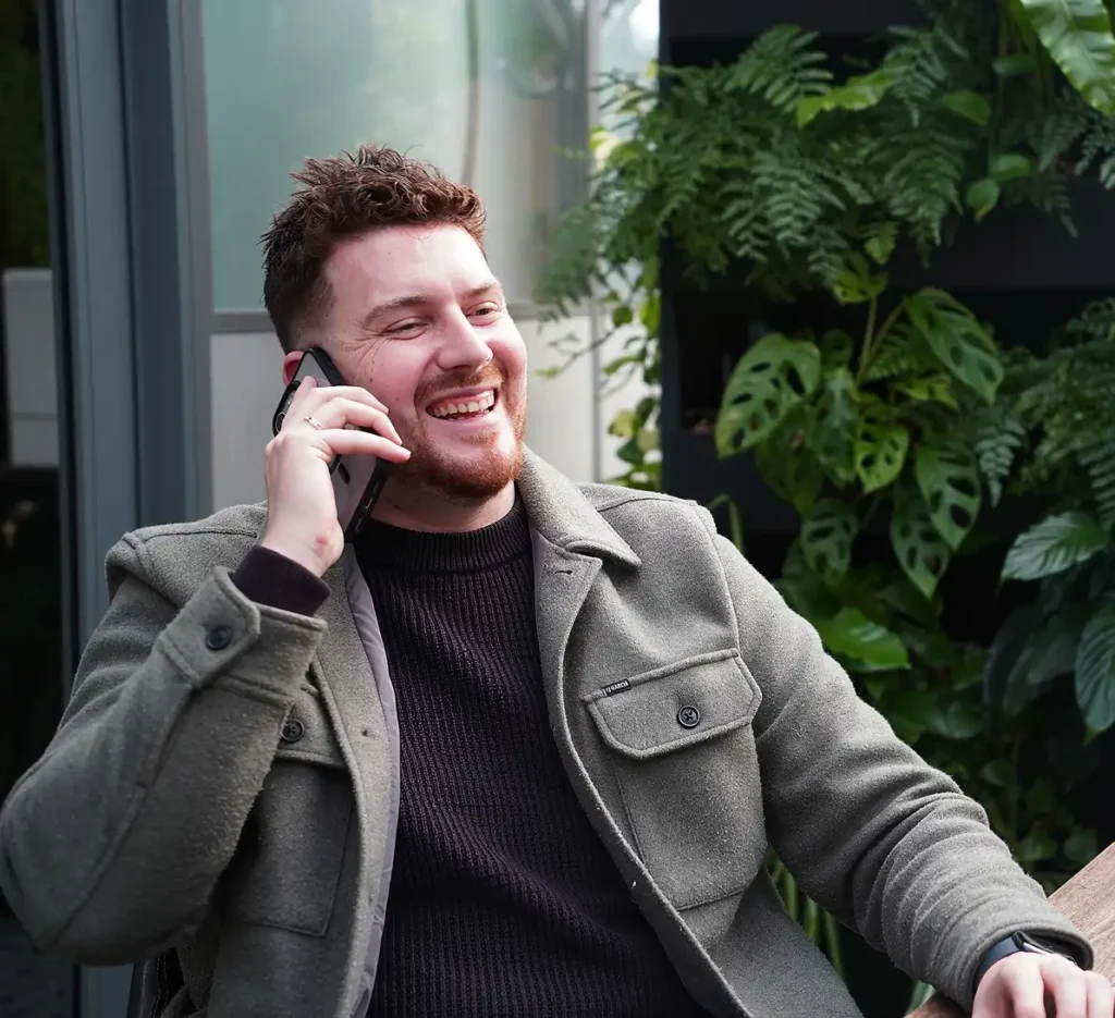 Kevin Flach lacht tijdens een telefoongesprek, met groene planten op de achtergrond.