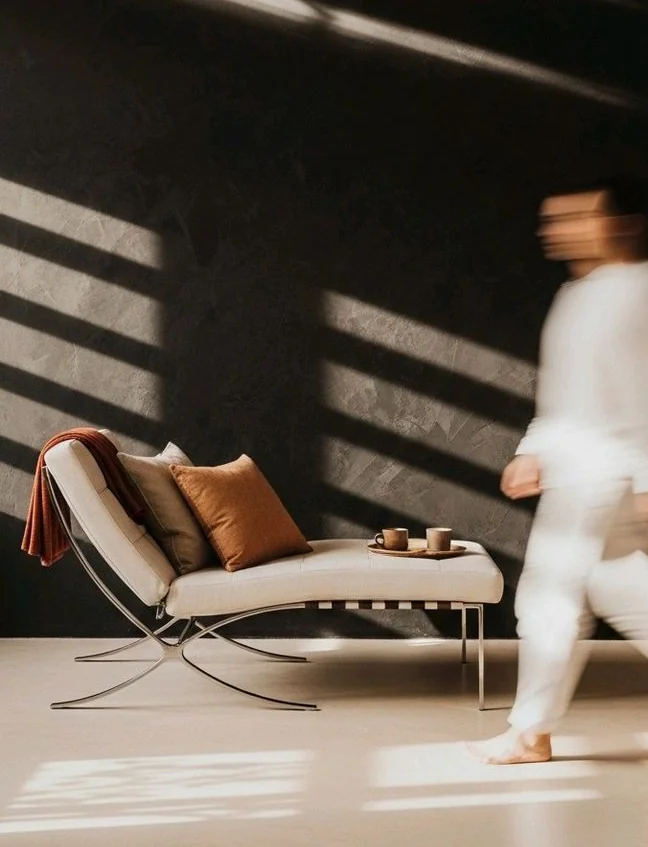 Een vrouw in witte kleding loopt door een zonovergoten interieur met een moderne stoel en warme kussens.
